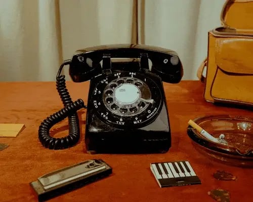 Vintage black rotary telephone with coiled cord on wooden desk, surrounded by small harmonica, cassette recorder, and brown leather bag
