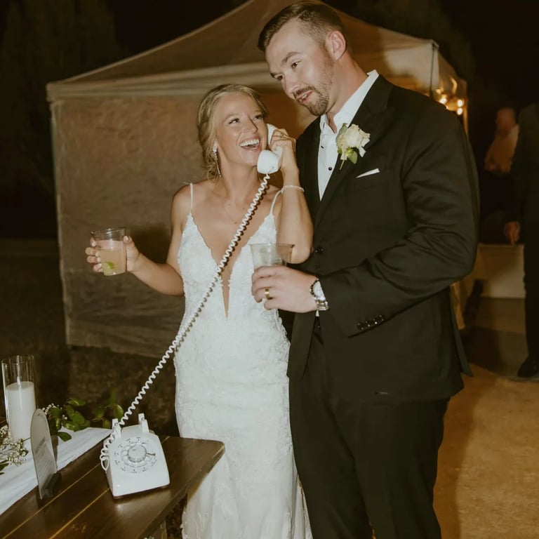 Bride and groom smiling while holding drinks at their wedding reception, standing near a vintage rotary phone and a tent backdrop