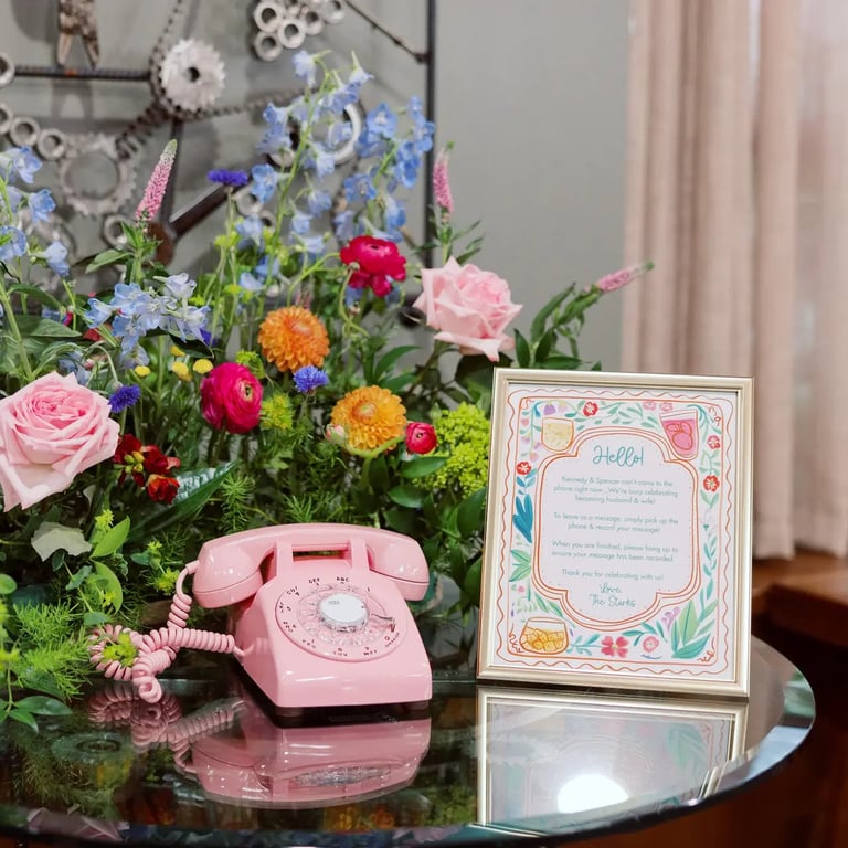 Vintage pink rotary phone next to colorful bouquet of roses, dahlias, and delphiniums with decorative card on glass table