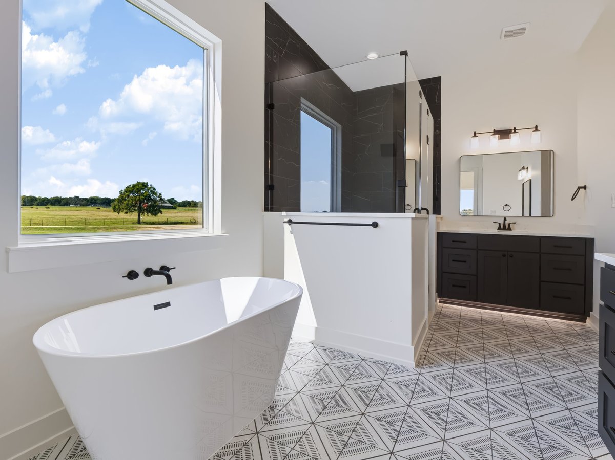 Modern luxury bathroom with freestanding white soaking tub, dark tile shower, double vanity, and large window overlooking pastoral landscape