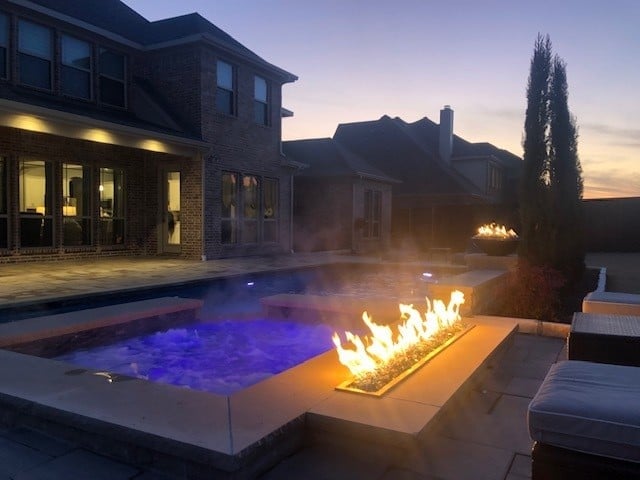 Luxury backyard at dusk featuring illuminated blue pool, linear fire pit, modern brick house, and manicured landscape with cypress tree