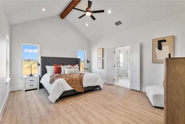 Spacious modern bedroom with gray upholstered bed, wood ceiling fan, white nightstands, wooden flooring, and abstract wall art