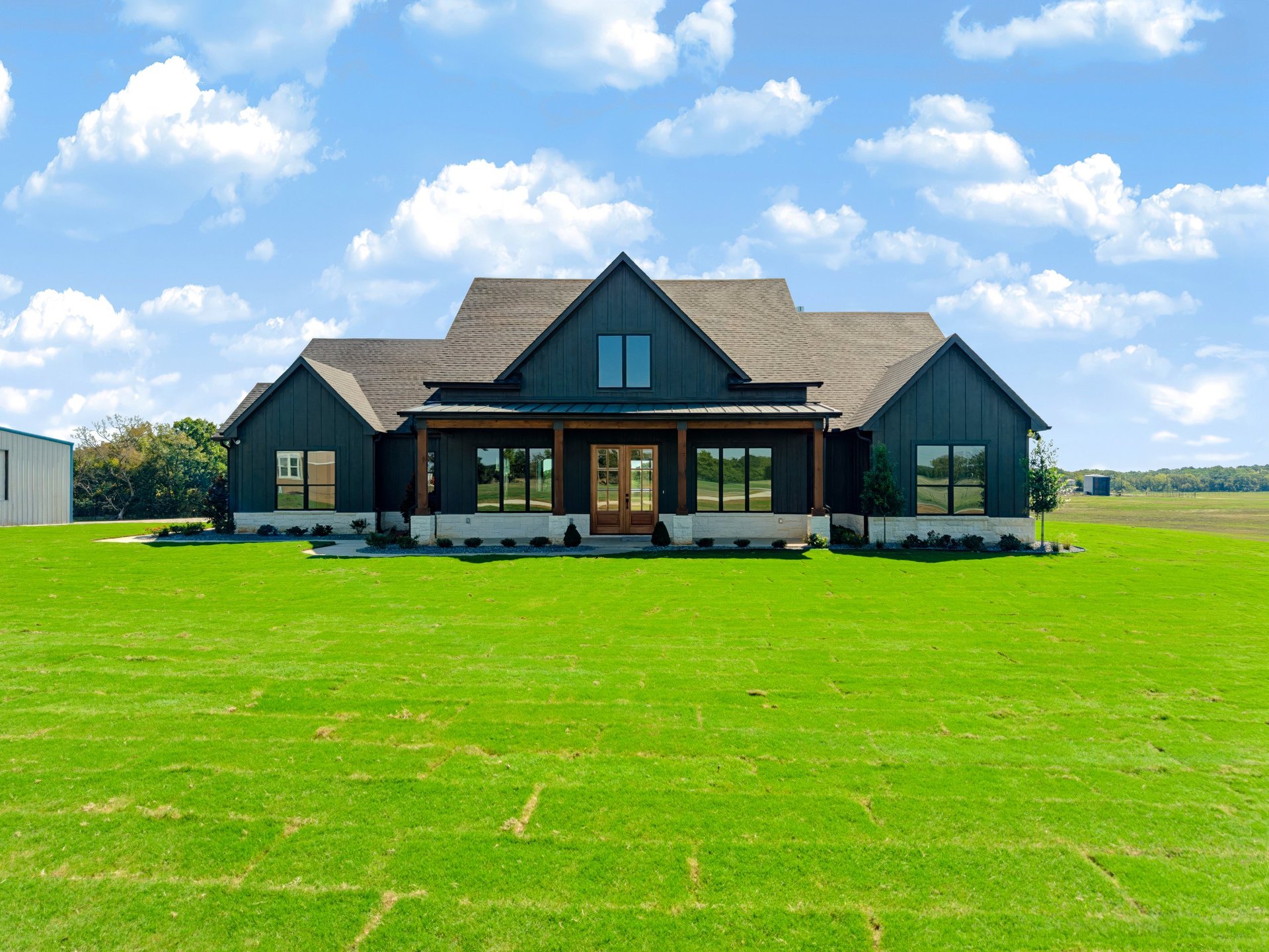 Modern black farmhouse with gabled roof set on expansive green lawn under blue sky with white clouds
