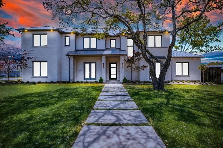 Modern two-story white house with gray roof, large tree in front yard, stone pathway leading to entrance, sunset sky background