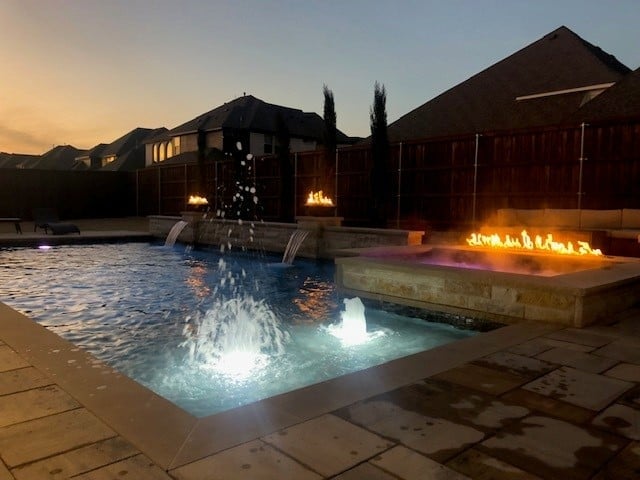 Luxury resort pool at dusk with illuminated fountain, fire pit, and modern buildings silhouetted against twilight sky
