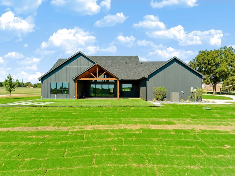 Rear elevation of the modern farmhouse custom home in Whitesboro, Texas — dark siding with a timber-framed gable and sliding glass doors opening to the backyard