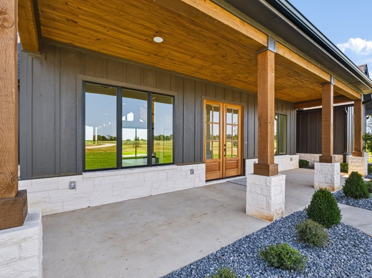 Modern home exterior with wooden beams, covered patio, concrete flooring, and landscaping overlooking open fields