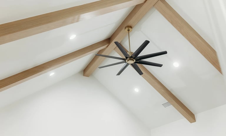 Modern black ceiling fan with brass accents mounted on white ceiling with wooden beams and recessed lights