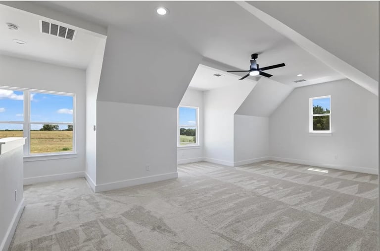 Bright modern attic bedroom with vaulted ceilings, skylights, large windows overlooking open fields, and a ceiling fan