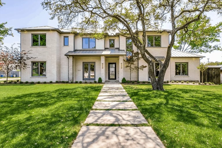 Modern two-story house with white stucco exterior, symmetrical facade, large tree in front yard, and stone walkway leading to the entrance