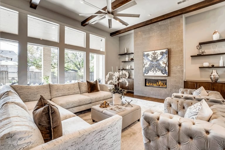 Spacious modern living room with neutral furniture, fireplace, large windows, ceiling fan, and tufted ottoman