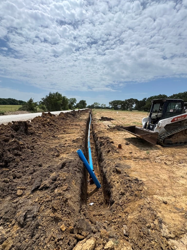 Construction site with blue pipe in trench, skid steer loader, and rural landscape under cloudy sky