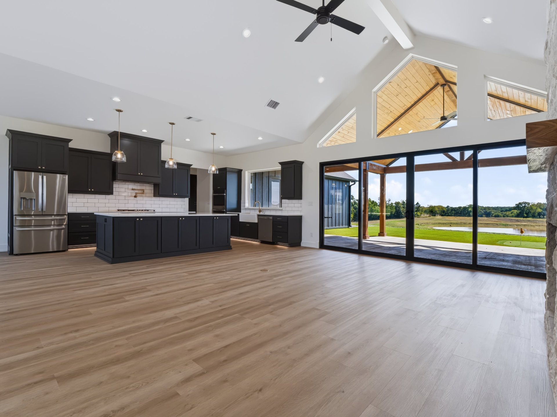 Modern luxury kitchen and open living space with large windows overlooking green fields, dark cabinets, stainless steel appliances, and wood flooring.