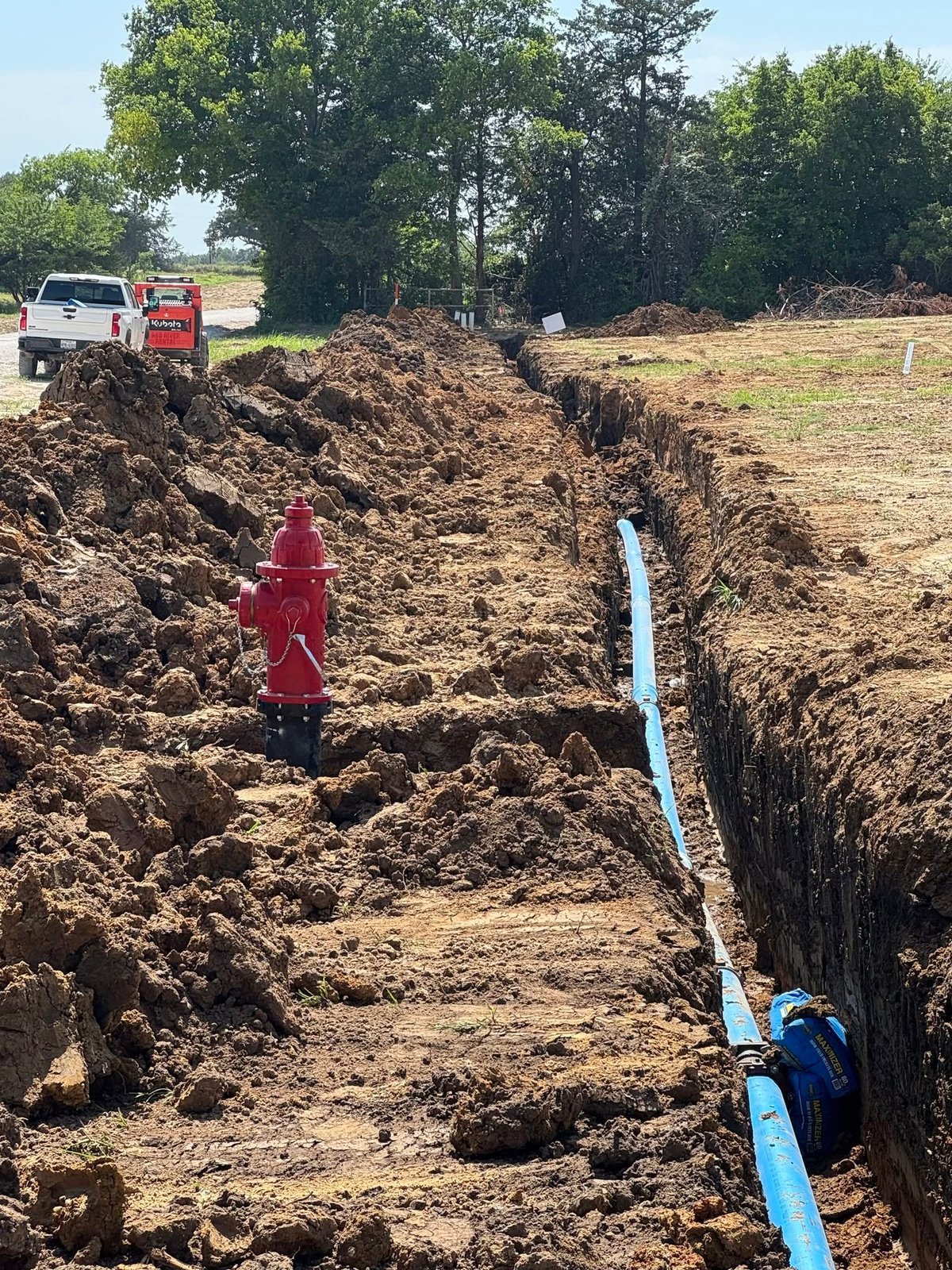 Construction site showing excavated trench with blue pipe installation, red fire hydrant, trucks, and trees in background