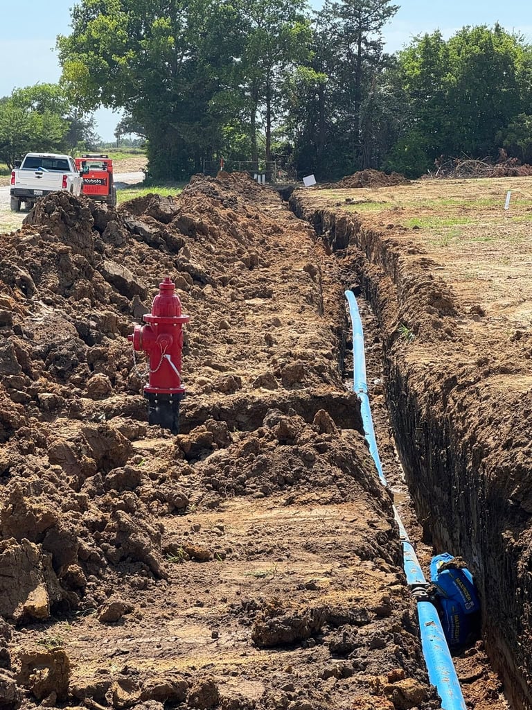 Construction site showing excavated trench with blue pipe installation, red fire hydrant, trucks, and trees in background