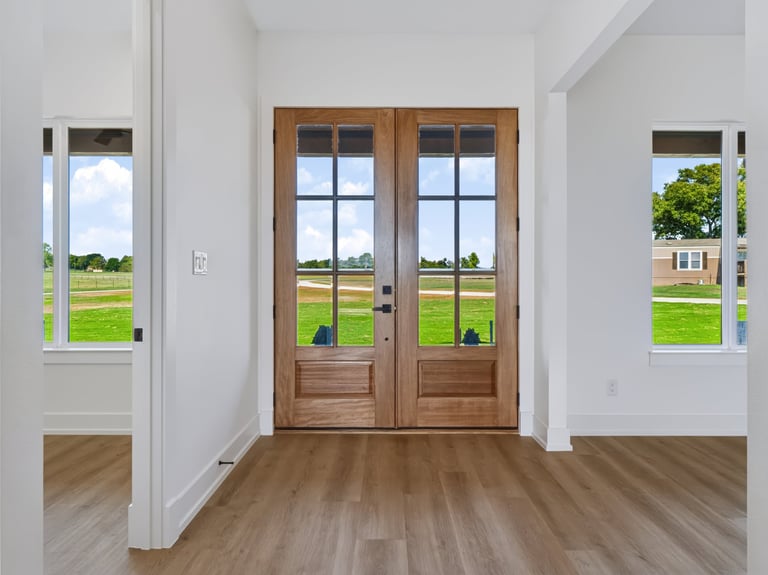 Bright interior hallway with wooden double doors, white walls, wood flooring, and countryside views through glass panels on either side.