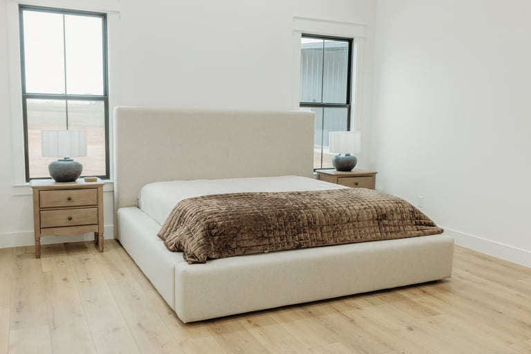 Bedroom in the completed Whitesboro custom home — cream upholstered bed with brown textured bedding, wooden nightstands and windows bringing in natural light