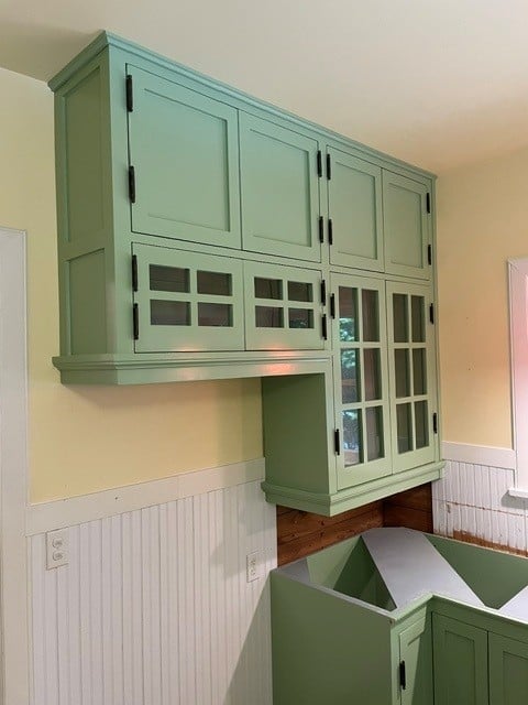 Sage green kitchen cabinetry with glass doors mounted on cream wall above lower cabinets with white beadboard trim