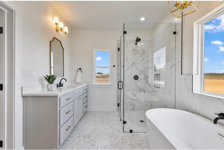 Modern luxury bathroom with marble tile shower, white freestanding tub, vanity sink with gray cabinetry, and windows overlooking countryside