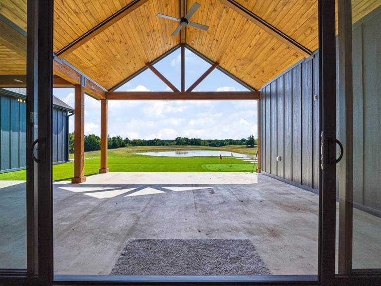 Modern covered patio with wooden beam ceiling and open doors overlooking a grassy field with trees