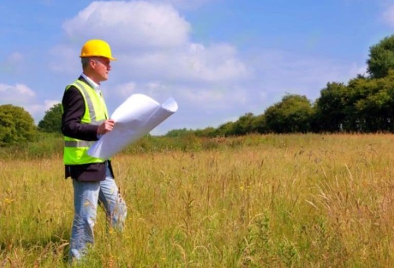 Construction worker in yellow hard hat and safety vest reviewing blueprints in open field under blue sky