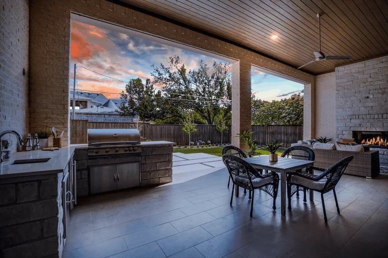Modern outdoor patio with built-in grill station, outdoor dining table, stone fireplace, and scenic sunset view overlooking a landscaped yard