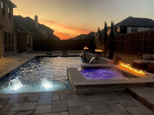 Backyard pool and hot tub with LED lighting at sunset, surrounded by houses and trees
