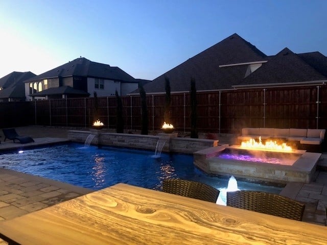 Backyard pool and fire pit at dusk with illuminated water features, wooden deck, and residential homes in background