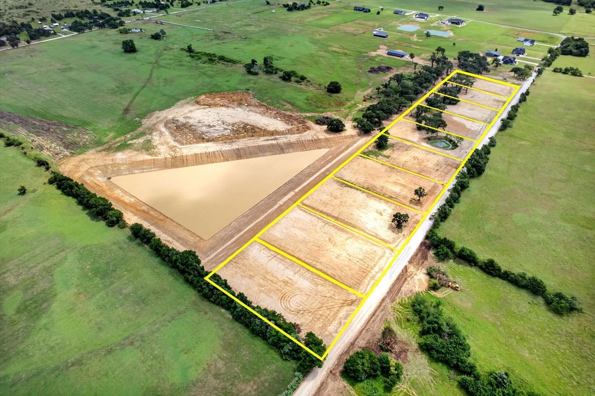 Aerial view of a construction or mining site with large earthen dam and subdivided land plots bordered by yellow markings, surrounded by green fields and scattered trees