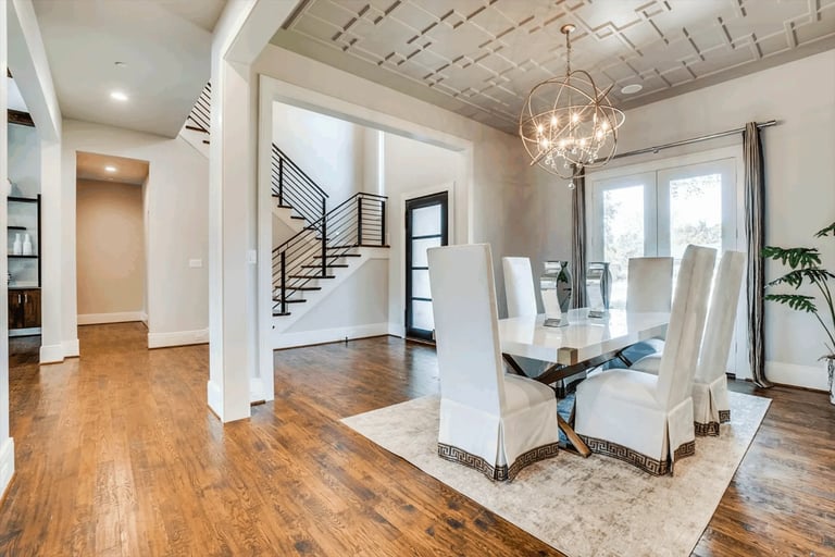 Modern open-concept home interior with wooden floors, white columns, staircase, dining area with upholstered chairs, and pendant lighting
