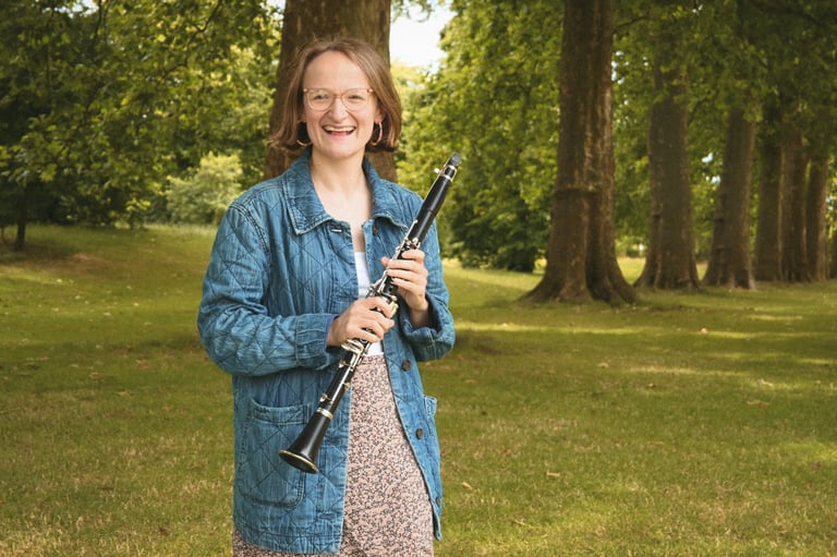 Smiling young woman holding a clarinet in a denim jacket outdoors in a tree-lined park