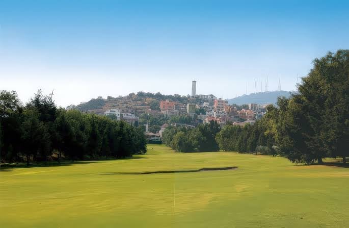 Green open field with trees framing the view and hillside town with buildings visible in the background under clear blue sky