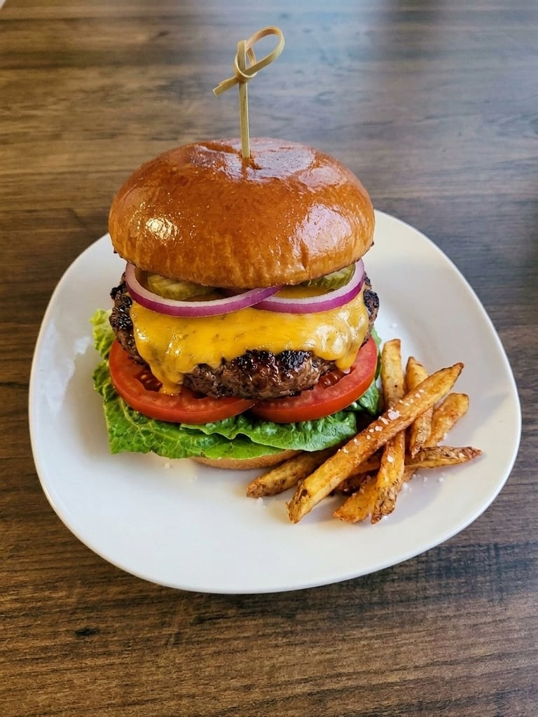 Gourmet cheeseburger with melted cheddar, tomato, red onion, and lettuce on a brioche bun with a gold pick, served with crispy fries on a white plate