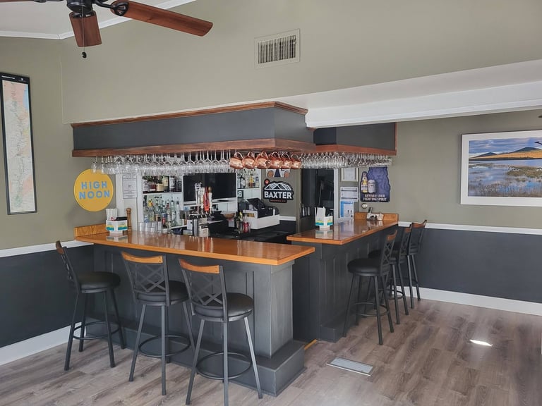 Modern home bar with dark wood counter, orange and black bar stools, shelving displaying bottles and barware, and industrial ceiling fan