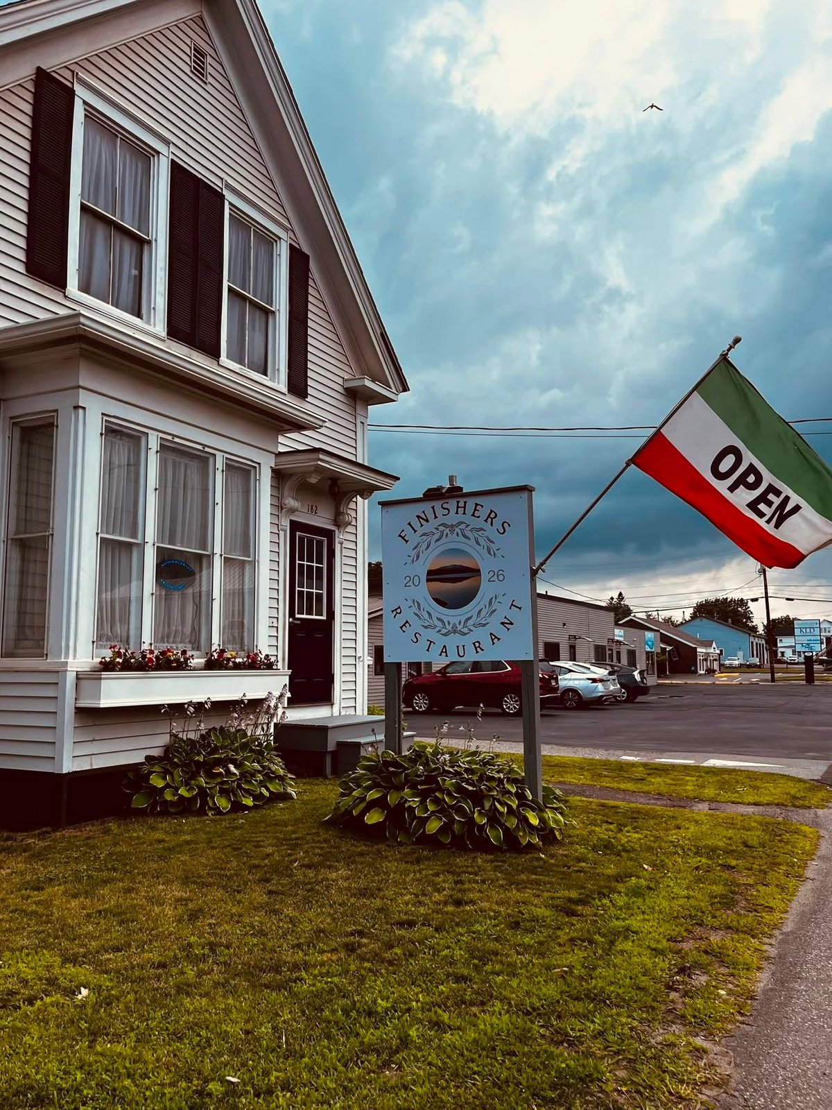 A white house with maroon shutters next to a restaurant sign with an open flag under a dramatic blue sky