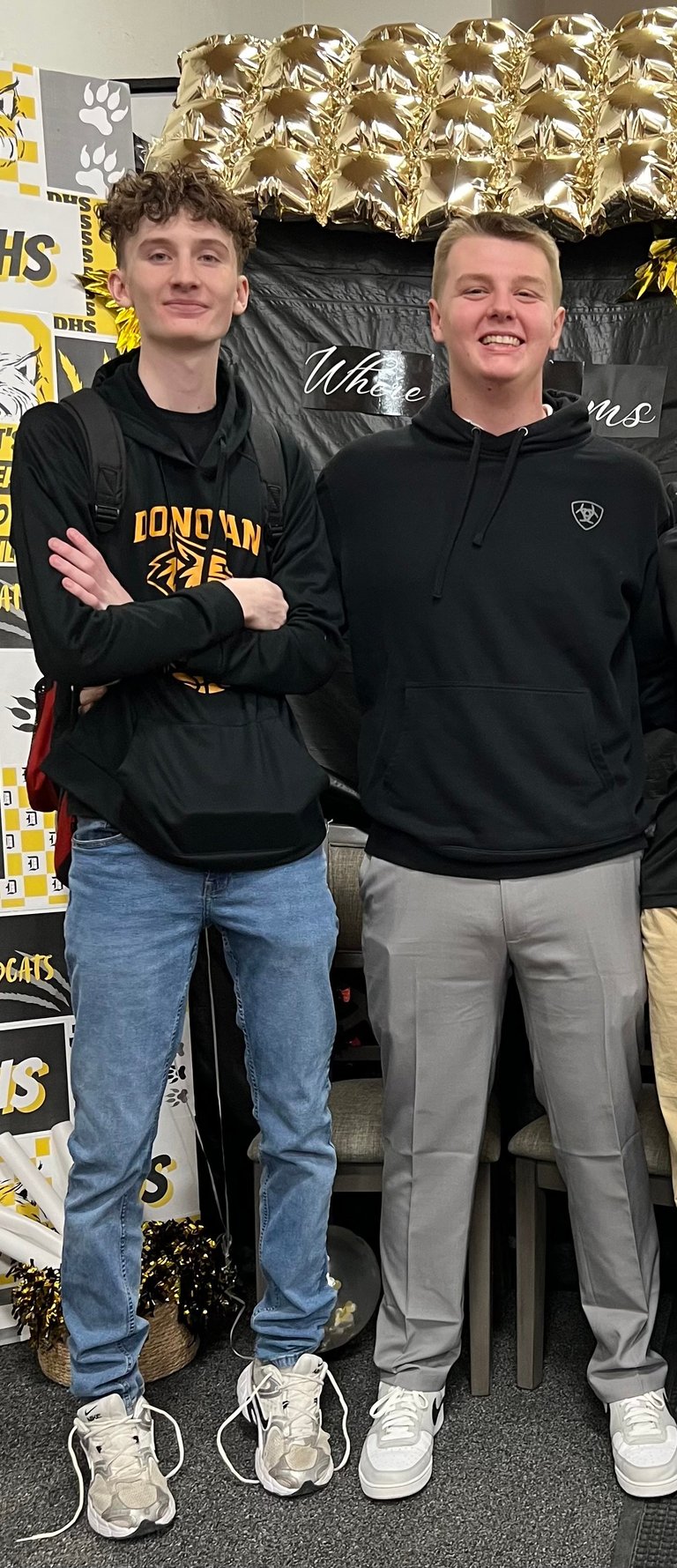Two young men standing side by side in front of a yellow and black school banner, wearing black hoodies and casual pants.
