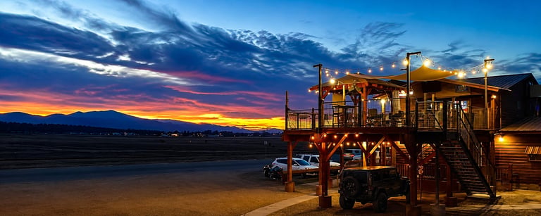 Illuminated wooden beach house at dusk with dramatic sunset sky and snow-capped mountains in the background