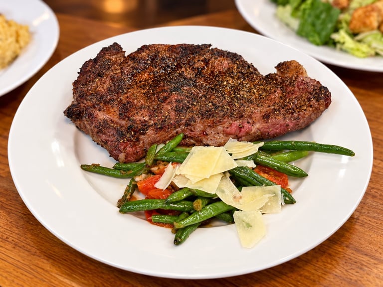 Crusted steak served on a white plate with green beans and shaved cheese, accompanied by side salads