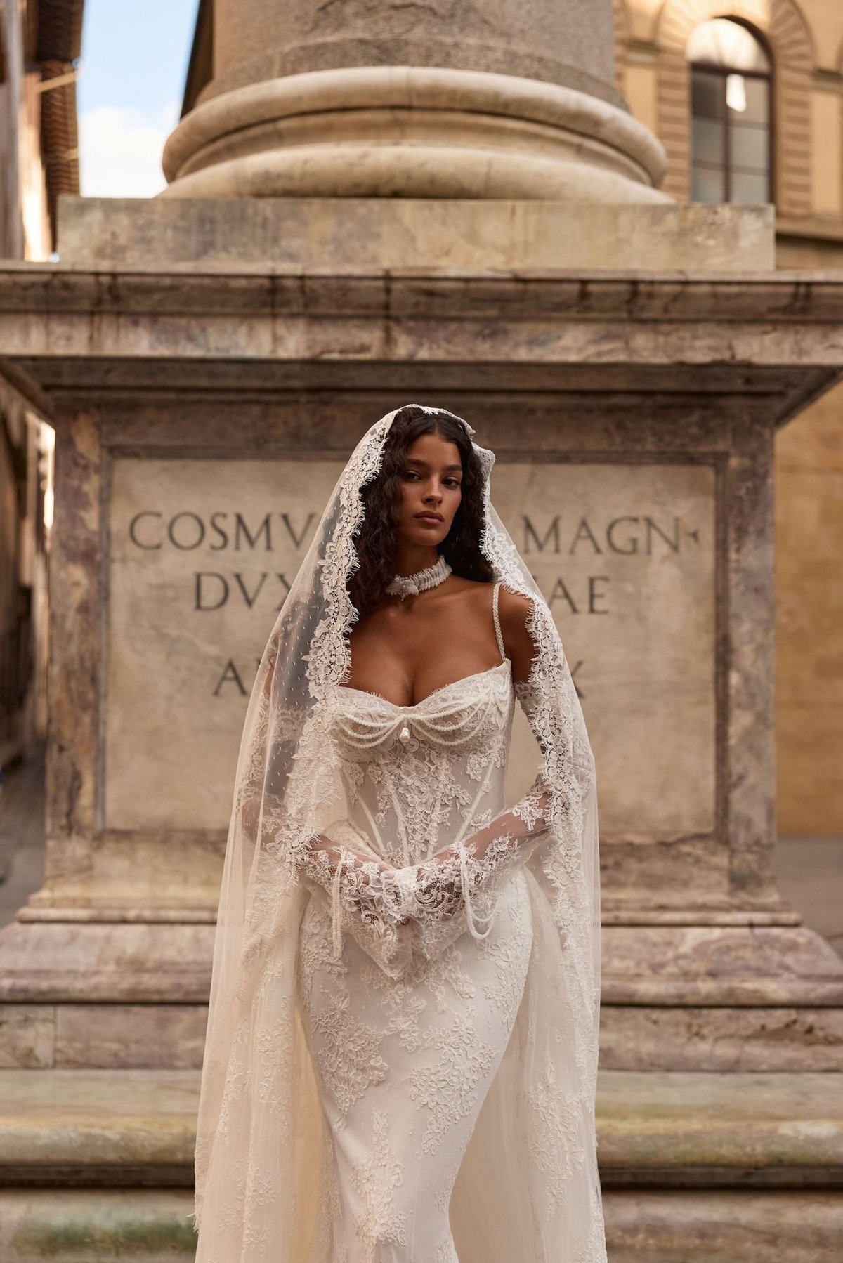 Woman in elegant ivory lace wedding dress with ornate veil standing before historic stone monument with classical inscriptions