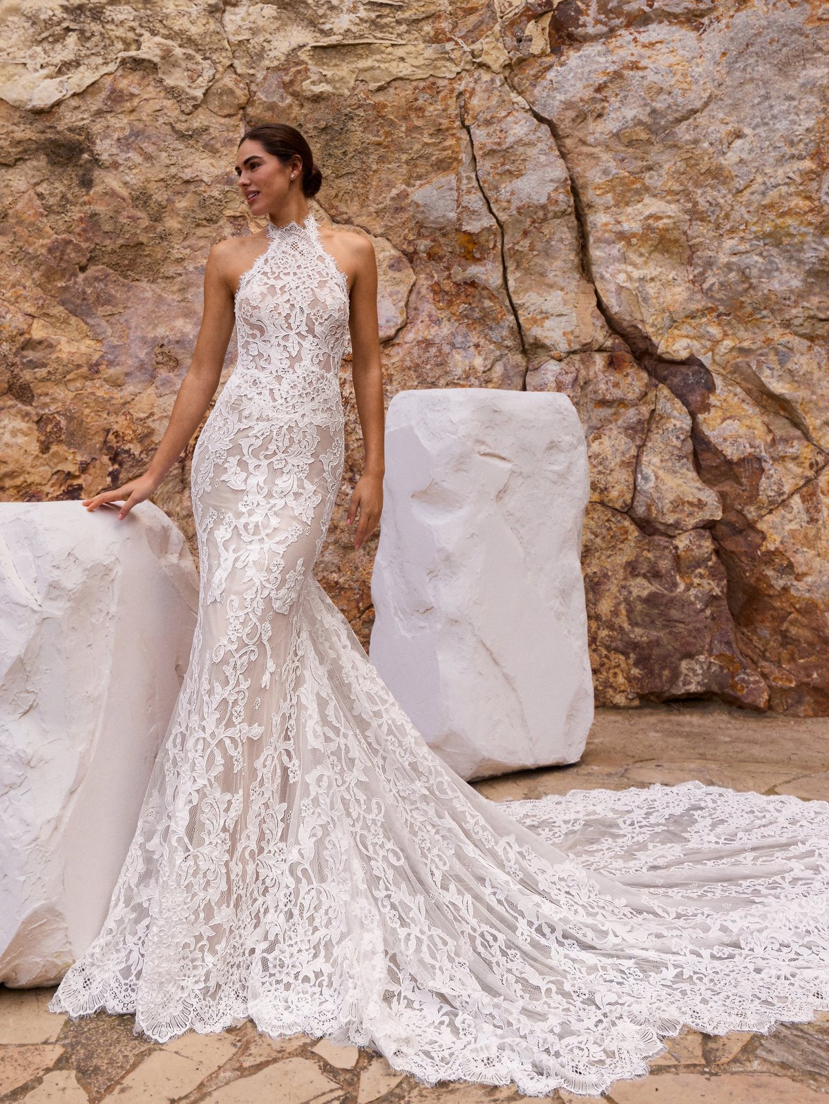 Bride in white lace mermaid wedding dress with long train posing against rustic stone wall backdrop