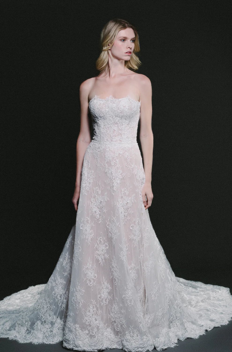 Woman modeling a strapless white lace wedding dress with a fitted bodice and flowing train against a dark background