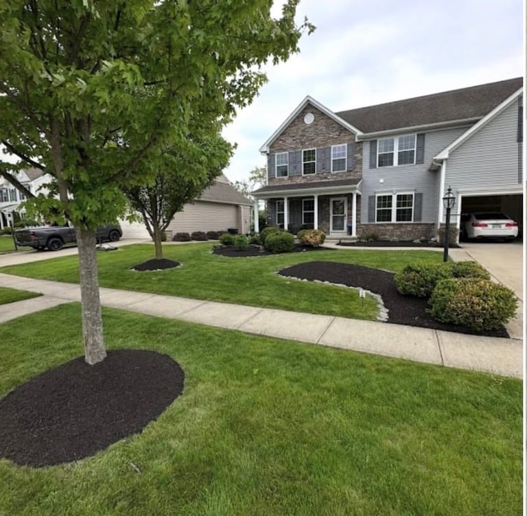 Two-story gray and stone house with attached garage, manicured lawn, mulch landscaping, and mature trees