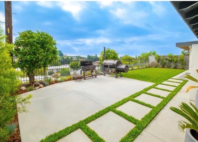 Modern backyard with concrete patio, stepping stones through green lawn, mature trees, and metal fence