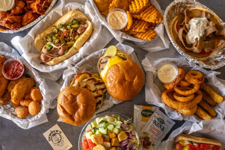 Overhead view of various fast food items including hot dogs, burgers, fried chicken, onion rings, and sides arranged on a gray surface