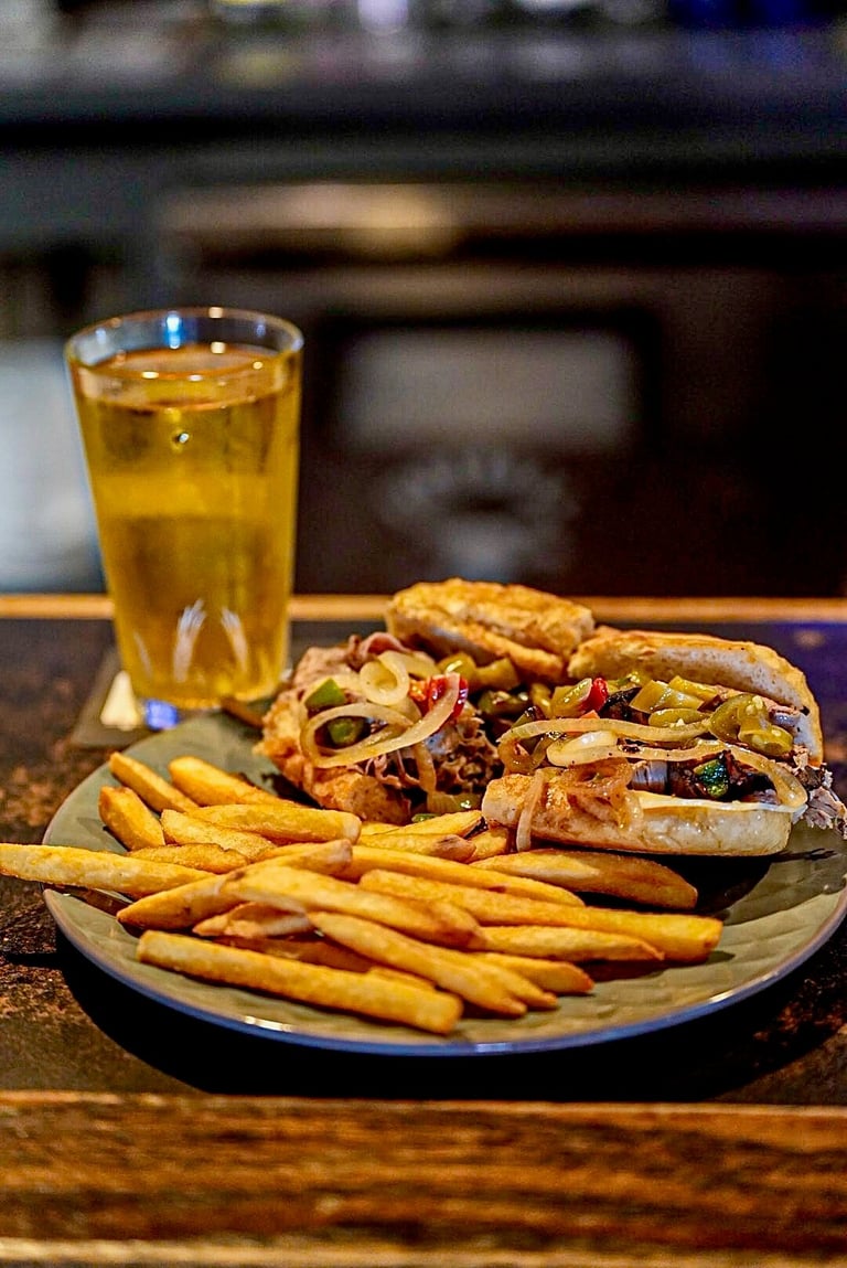 A plate of food with two sandwiches, french fries, and a glass of beer on a wooden table in a bar setting