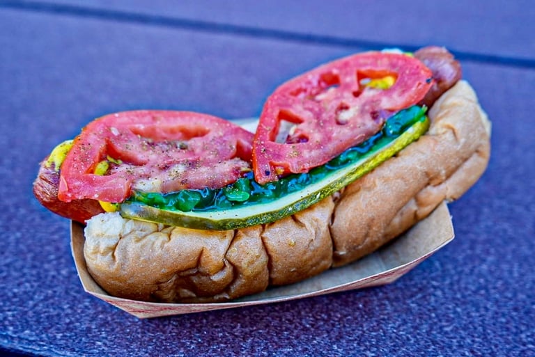 Colorful hot dog with pink frosting, yellow mustard drizzle, and green icing on a toasted bun, served on a blue speckled surface