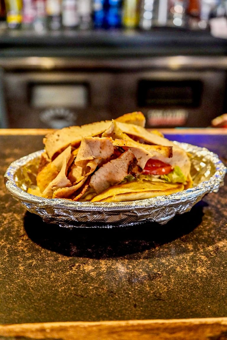 A taco with sliced meat, lettuce, and tomato served in a foil container with crispy tortilla chips on a bar counter.