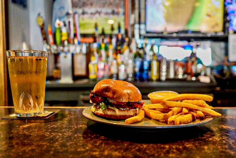 A burger and fries served on a bar counter with a glass of cola, blurred bar bottles in the background