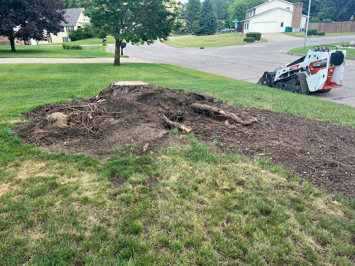 Pile of mulch and soil on residential lawn with skid steer loader and houses in background