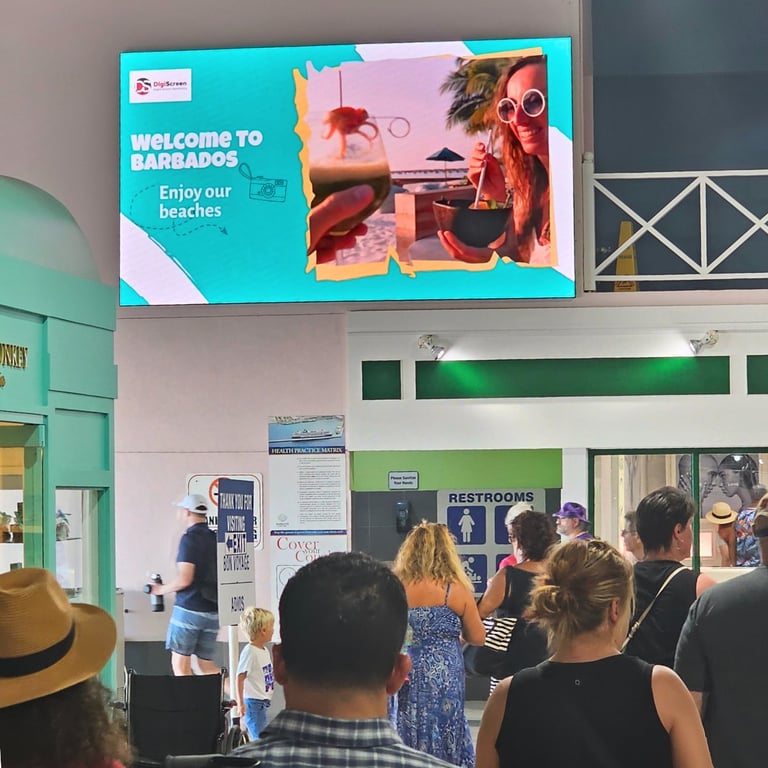 Digital display screen showing Welcome to Barbados tourism ad above crowded indoor venue with visitors and restroom signage